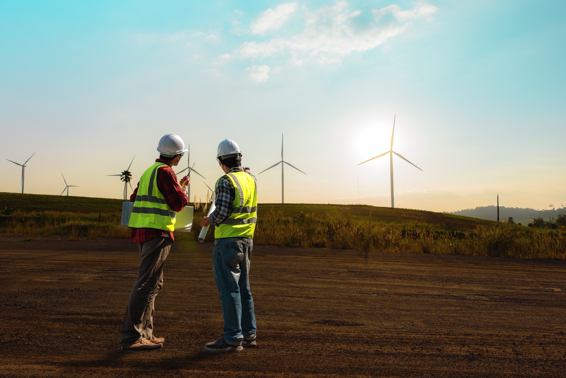 Two maintenance engineer working on wind turbine farm in sunset, discussed plan for the maintenance of wind turbines while holding blueprint of layout plan.